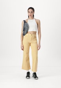 Pantaloni a gamba larga in corduroy giallo, canotta corta bianca, borsa a tracolla in denim, scarpe nere con motivo testurizzato, modello in piedi su sfondo bianco.