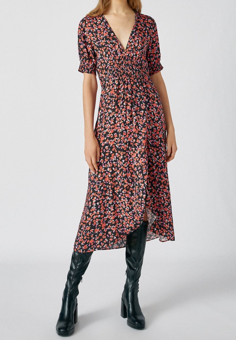 Vestido floral en negro, con un escote en V profundo, mangas cortas abullonadas y una silueta fluida con una sutil abertura frontal. Combinado con botas negras hasta la rodilla.