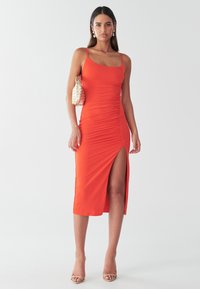 Czerwona obcisła sukienka na ramiączkach, z bocznym rozcięciem i marszczoną fakturą. W zestawieniu ze złotą mini torebką i beżowymi sandałami na paski.