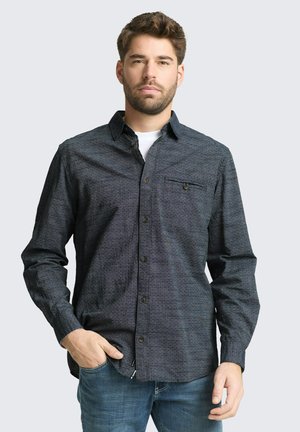 Mann mit Bart, der ein dunkel gemustertes Hemd über einem weißen T-Shirt trägt, steht mit einer Hand in der Tasche einer blauen Jeans vor schlichtem Hintergrund.