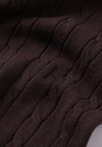 Καφέ ύφασμα με πλεκτό σχέδιο, που παρουσιάζει ανάγλυφα μοτίβα, με υφή στην επιφάνεια και ένα διακριτικό λογότυπο κεντημένο σε πιο σκούρα απόχρωση.