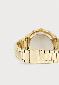 Gullfarget metallklokke med lenkearmbånd. Rund klokkege Case med glassbakside. Har en krone og chronograf-knapper på siden.
