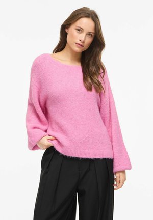 Femme aux longs cheveux bruns portant un pull rose ample et duveteux et un pantalon noir plissé, debout avec une main dans la poche.