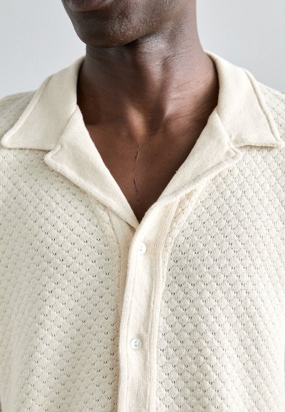 Gebreide cardigan in crèmekleur, met een ruitpatroon, knoopsluiting aan de voorkant en een opgevouwen kraag. Zachte textuur met zichtbare steekdetails.