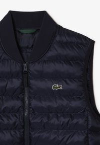 Tmavě modrá zateplená vesta s vodorovným prošíváním, žebrovaným límcem, předním zapínáním na zip a vyšívaným zeleným krokodýlím logem.