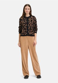 Egy nő áll, mosolyogva, fekete és bézs leopárdmintás kardigánt, bézs kordbársony nadrágot és fekete cipőt viselve egy sima háttér előtt.