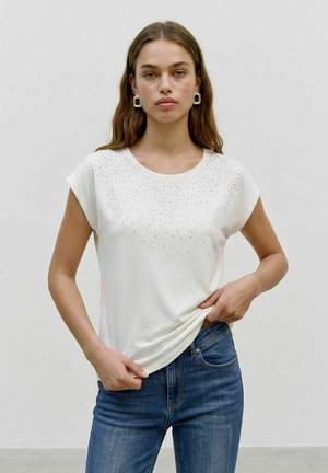 Ung kvinde iført en hvid nittebesat kortærmet top og blå jeans, stående mod en ensfarvet lys baggrund.
