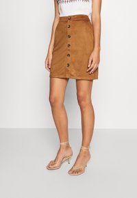 Brun mocka A-linjeformad kjol med hög midja, framknappstängning och sömnadsdetaljer. Matchas med gyllene högklackade sandaler med remmar.