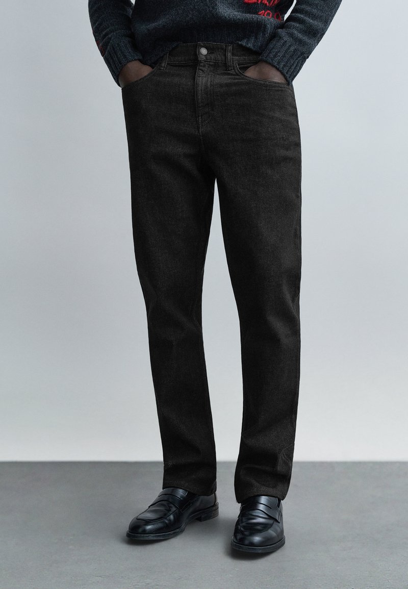 Černé džíny z denimu s rovnými nohavicemi a jemnou texturou. Jsou kombinovány s černými společenskými botami a šedým svetrem.