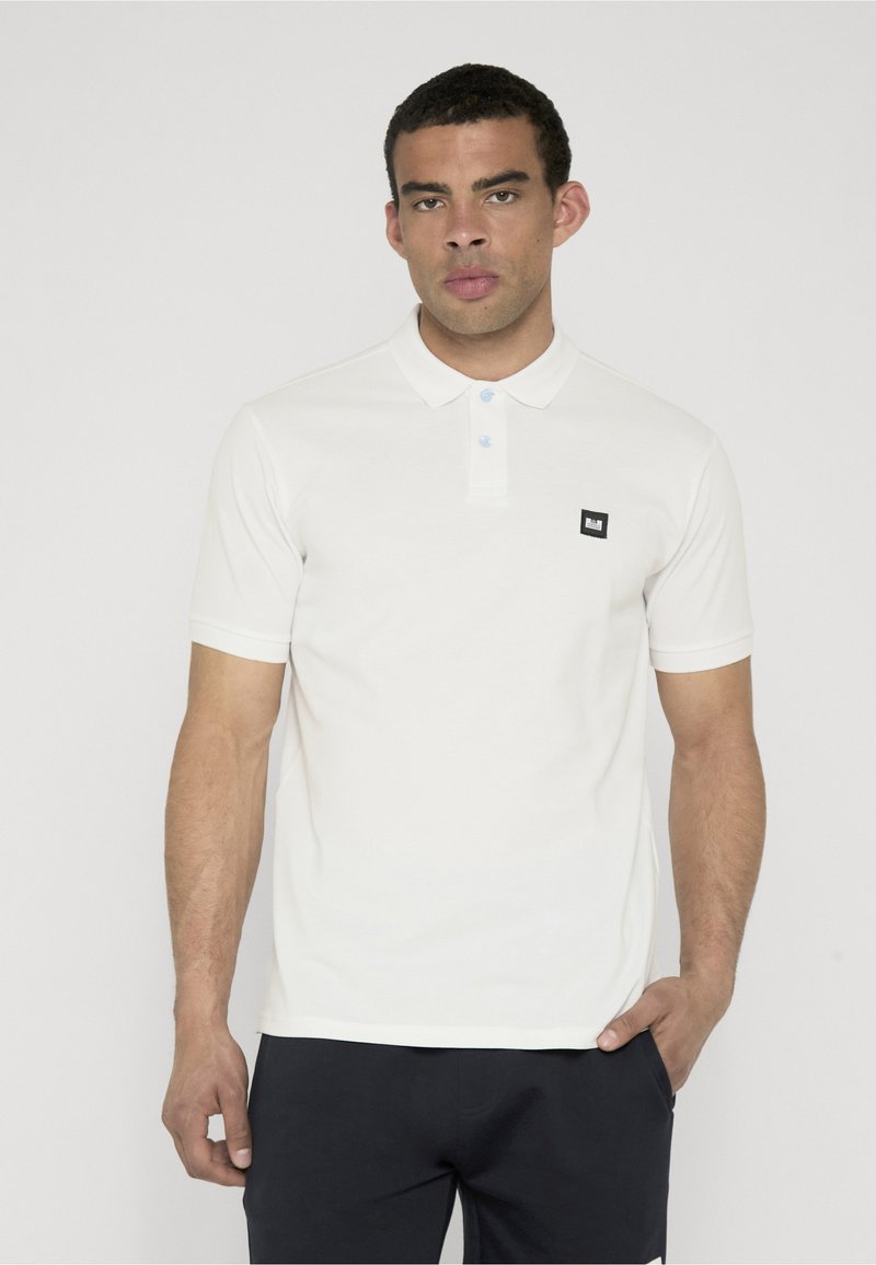 Homme aux cheveux courts portant un polo blanc à manches courtes et un pantalon noir, debout avec une main dans la poche devant un fond uni.