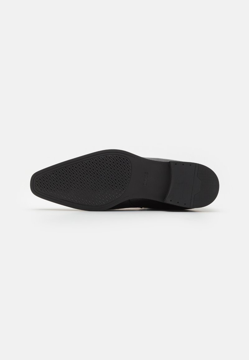 Semelle de chaussure noire avec une surface texturée, présentant une forme pointue et un motif de semelle distinct pour l'adhérence. Design minimaliste.