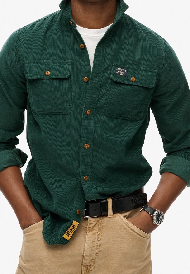 Camicia in velluto a coste verde scuro con due tasche sul petto, bottoni marroni e patch con logo. Indossata con cintura nera e pantaloni beige. Orologio argento visibile.