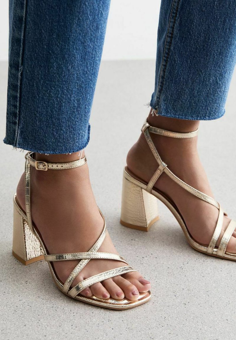 Gouden bandjes sandalen met een vierkante neus, blokhak en textuuraccenten, gedragen met donkerblauwe cropped jeans op een lichte ondergrond.