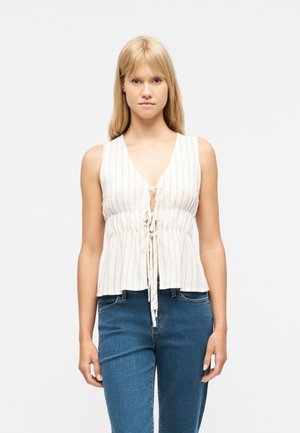 Donna bionda con capelli lunghi che indossa un top bianco senza maniche a righe con laccetti sul davanti e jeans di denim blu.