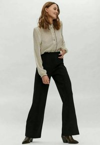 Blusa clara com pequenas bolinhas, mangas longas e botões; calças largas de cintura alta pretas; botas de salto castanhas.