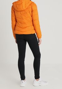Orange vadderad jacka med huva, med quiltade detaljer och elastiska muddar, bärs över svarta skinny jeans och vita sneakers.