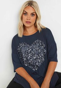 Marinblå t-shirt i mjukt tyg med ett stort hjärtformigt mönster av paljetter i silver och mörkblått. Kort ärm och avslappnad passform.