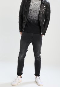 Persona che indossa una giacca di pelle nera, felpa con grafica di teschi, jeans neri risvoltati alle caviglie e sneaker bianche, in piedi con le mani nelle tasche.