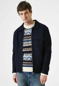 Man in een marineblauwe zip-up jack over een gebreide trui met patronen in blauw, wit en bruin, gecombineerd met blauwe spijkerbroek, staand tegen een witte achtergrond.