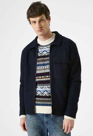 Man in een marineblauwe zip-up jack over een gebreide trui met patronen in blauw, wit en bruin, gecombineerd met blauwe spijkerbroek, staand tegen een witte achtergrond.