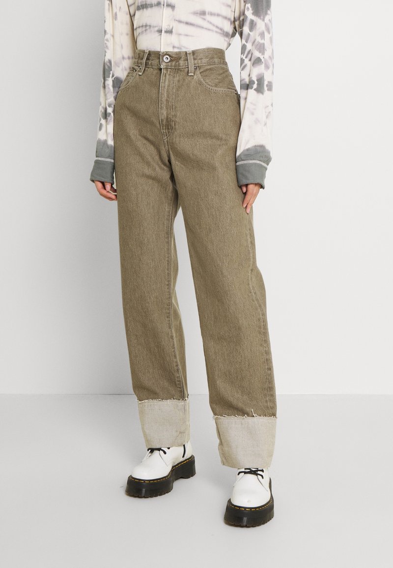Jean large kaki avec des revers gris clair effilochés, en denim. Porté avec un t-shirt à manches longues tie-dye et des chaussures blanches épaisses.