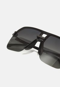 Occhiali da sole neri oversize con design a lente piatta, caratterizzati da una forma angolare audace e un sottile ponticello in metallo che collega le lenti.