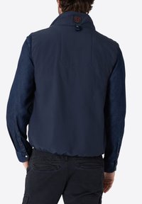 Dunkelblauer gepolsterter West mit hohem Kragen, der ein gesticktes Logo auf der Rückseite und ein Denim-Hemd mit langen Ärmeln darunter hat.