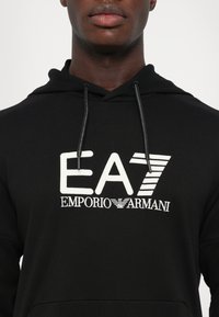 Sweat à capuche noir avec logo blanc EA7 Emporio Armani sur la poitrine, porté par une personne montrée du cou au milieu du torse.