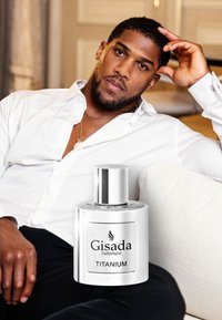 Gisada Titanium perfume bottle with a silver cap and label, next to a man in a white shirt, seated on a light-colored sofa.