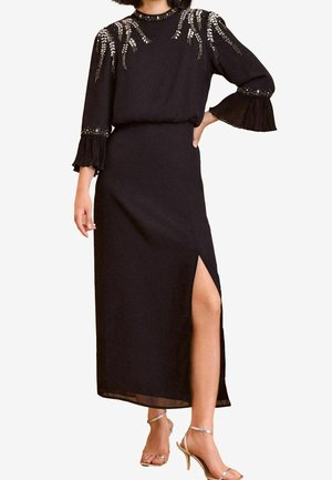 Robe midi noire avec épaules et manches ornées, manches plissées 3/4, fente latérale à la cuisse, portée avec des talons métalliques à bride cheville.