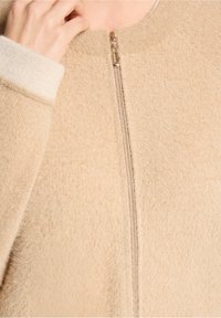 Veste beige à fermeture éclair avec un tissu doux et texturé. Elle présente un col montant et des accents de poignets de couleur claire, complétés par une fermeture éclair en métal.