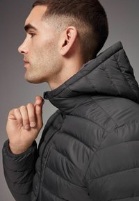 Jachetă puf din material negru, cu un model cusut, guler cu fermoar și glugă, prezentată pe o persoană care își ajustează fermoarul, pe un fundal neutru.