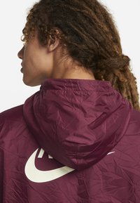 Temno rdeča jopa s kapuco iz lahkega, zmečkanega blaga. Na hrbtu ima velik bež Nikejev logotip, kapuca pa je tesno prilegajoča.