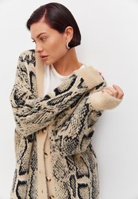 Beige neulepusero mustalla käärmeennahkakuviolla, pörröinen rakenne, löysä malli ja suuret napit. Malli on kietoutunut neuletakkiin, ja hänellä on suuret hopeiset renkaat korvissa.