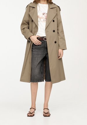 Femme portant un trench-coat beige sur un T-shirt blanc imprimé de chevaux, un short en jean noir à hauteur des genoux, une ceinture marron et des sandales marron à lanières.