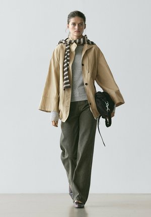 Kobieta idąca, ubrana w beżową kurtkę, szary sweter, pasiasty szalik, ciemne spodnie o szerokich nogawkach, trzymająca czarną torbę na neutralnym tle.