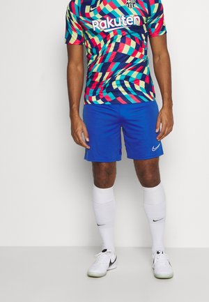 Mann trägt ein mehrfarbiges Fußballtrikot mit geometrischem Muster, blaue Shorts, weiße hohe Socken und weiße Nike-Fußballschuhe, steht vor einem einfarbigen Hintergrund.