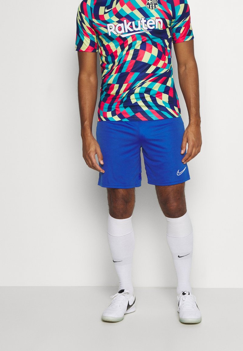 Homme portant un maillot de football multicolore à motif géométrique, un short bleu, des chaussettes hautes blanches et des crampons Nike blancs, debout devant un fond uni.