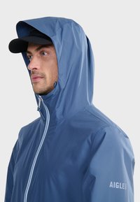 Blue waterproof jacket with a zip closure, adjustable hood, and branding on the sleeve, made of smooth, lightweight material.