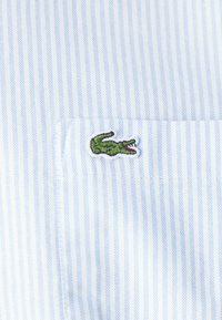Lichtblauw gestreepte stof van een overhemd met een klein groen krokodillenlogo geborduurd op de borstzak. Subtiele textuur van de strepen.