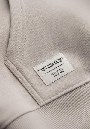 Jasno beżowa tkanina z haftowanym dekoltem i teksturowanym prążkowanym wykończeniem. Zawiera wplecioną metkę z czarnym tekstem i logo.