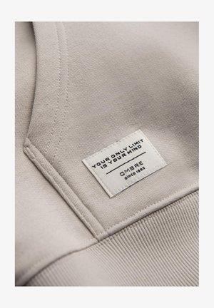 Jasno beżowa tkanina z haftowanym dekoltem i teksturowanym prążkowanym wykończeniem. Zawiera wplecioną metkę z czarnym tekstem i logo.