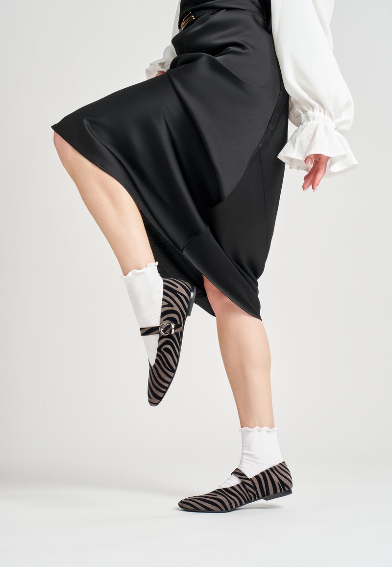 Zapatos planos a rayas en negro y marrón con puntera puntiaguda, una hebilla decorativa, y combinados con calcetines tobilleros de canalé blancos.