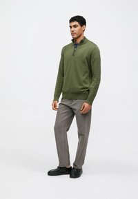 Groene pullover trui met een halve rits kraag, gecombineerd met lichtgrijze broek en zwarte schoenen. Gladde stof en nauwsluitend ontwerp.