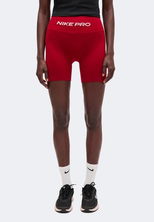 Άτομο που φοράει κόκκινα σορτς Nike Pro, μαύρο τοπ, λευκές κάλτσες Nike και μαύρα αθλητικά παπούτσια Nike, στέκεται μπροστά σε ένα μονόχρωμο φόντο.