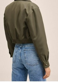 Graue, olivgrüne Jacke mit elastischem Saum und knopfverschlossenen Ärmeln, kombiniert mit hochgeschnittenen hellblauen Jeans mit Gesäßtaschen.