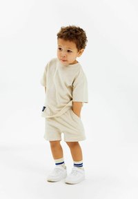 Beige bomullströja och shorts-set, avslappnad passform med korta ärmar, i kombination med vita sneakers och randiga strumpor, stående framför en vit bakgrund.