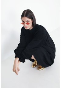 Zwarte langmouw jurk met geplooide manchetten, gedragen met metallic gouden schoenen. Model met rode hexagonale zonnebril, zittend op de vloer.