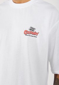 Hvid bomulds t-shirt med korte ærmer, der har et flerfarvet grafisk print, inklusive "Feelin' Fresh!" og "Groovin' with the Fruit Crew."