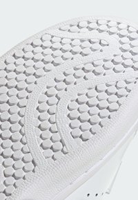 Baltā sneaker zole ar teksturētu modeli, kas sastāv no maziem apļiem un viļņveida līnijām. Malas ir gludas ar nelielu spīdumu.